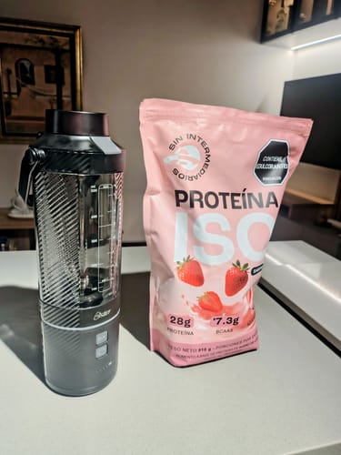 Cliente muestra la bolsa rosa de Proteína ISO Fresa junto a una licuadora portátil en un mostrador.