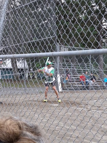 image 2 out of 2: Partial view of a reviewer at bat wearing the custom white and green crocodile baseball jersey, seen through a fence.
