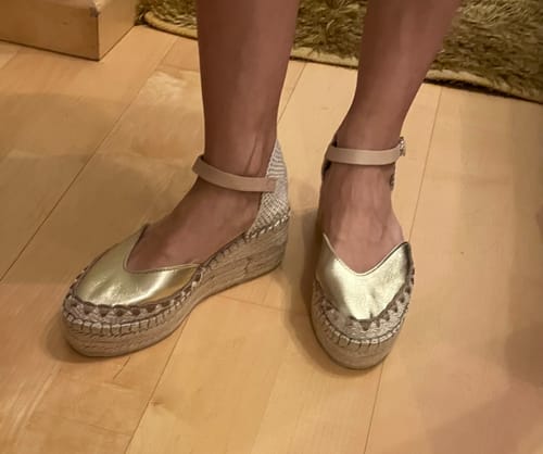 Cliente mostrando sus Alpargatas Lía Oro doradas en un suelo de madera, vista frontal.