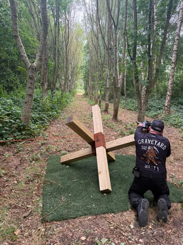 Reviewer wearing the black Undertaker Long Sleeve Jersey while kneeling and aiming a firearm in a wooded area.