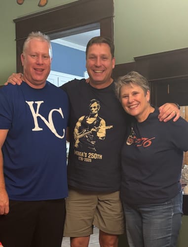 Customer's photo of a person wearing the dark Lincoln 250 T-Shirt standing between two smiling people.
