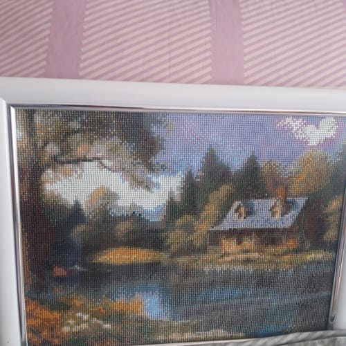 Kunde zeigt das fertige und gerahmte Diamond Painting 'Hütte im Wald' in einem weißen Rahmen.