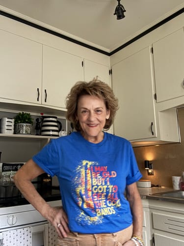 Reviewer smiling in a kitchen while wearing the royal blue "I May Be Old" graphic T-shirt.