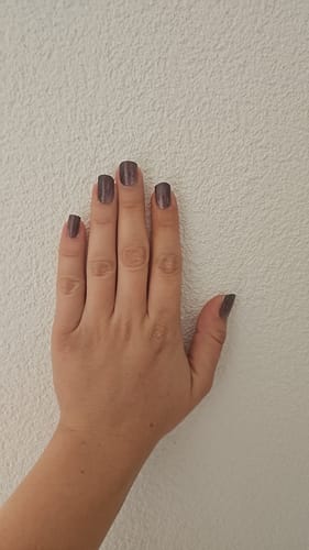 Hand einer Kundin, die die glitzernden Crystal Merlot UV-Gel-Nagelfolien auf kurzen Nägeln vor einer weißen Wand zeigt.
