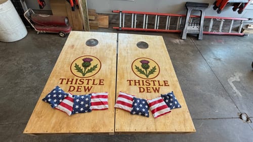 Customer's custom cornhole board stickers with a thistle logo and 'Thistle Dew' text applied to two wooden boards.