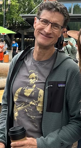 Reviewer smiling while wearing the grey Lincoln Guitar T-Shirt under an open zip-up hoodie.