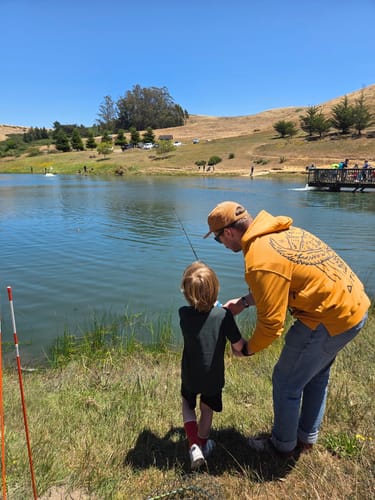 Reviewer wearing the yellow Trailhood hoodie while helping a child fish by a lake on a sunny day.