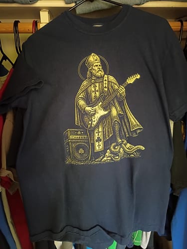 Customer's dark blue Saint Patrick Guitar T-Shirt on a hanger, showing the full front graphic of the saint playing a guitar.