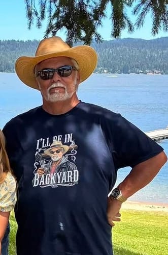 A reviewer wearing the dark custom 'I'll Be In The Backyard' t-shirt while standing outdoors by a lake.