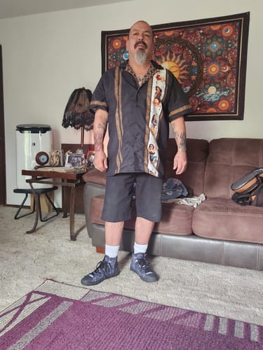 Customer standing in a living room wearing the black Chicana Pinups Guayabera shirt and shorts.