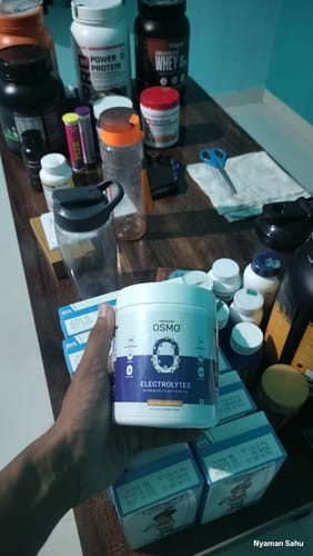 Reviewer holding a white tub of OSMO Electrolyte Hydration Blend in front of a table with other fitness supplements.
