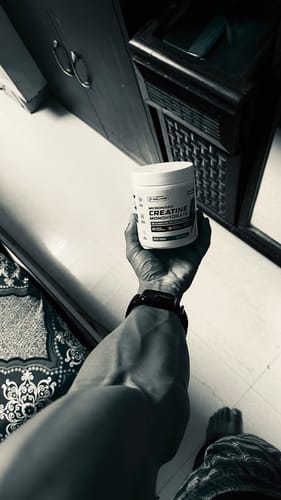 Reviewer's black and white photo showing a muscular arm holding a tub of Wellcore Micronised Creatine Monohydrate.