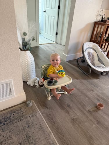 Customer's happy baby sitting in the checkered Parker portable booster chair V2 on a floor at home.