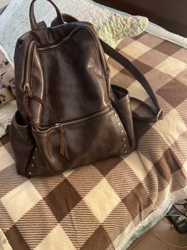 Cliente muestra su Mochila Tachas Puller Gamuza Boho de color marrón, con tachas y cierres, sobre una cama con una manta a cuadros.