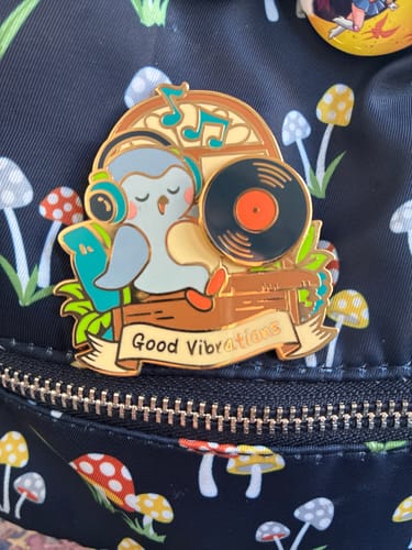 Customer's Good Vibrations enamel pin attached to a black backpack with a colorful mushroom pattern.