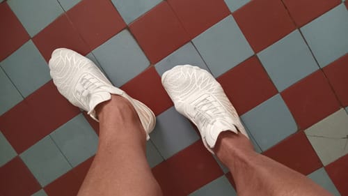 Reviewer's point-of-view looking down at their white Flexies Barefoot Shoes on a checkered tile floor.