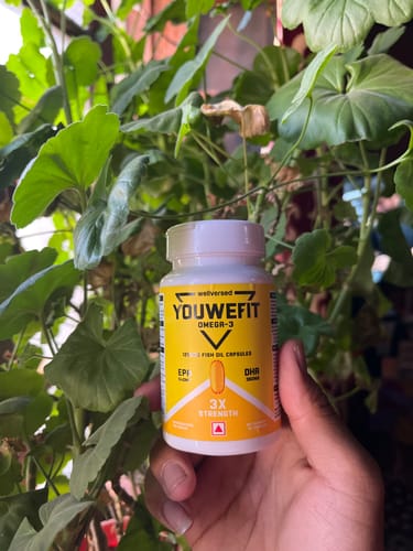 Customer holds a bottle of YouWeFit Omega-3 Fish Oil in their hand, with a leafy green plant in the background.