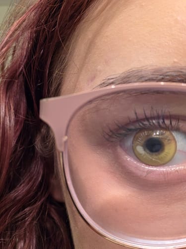 Close-up of a reviewer's eye wearing the yellow prescription colored contact lens, viewed behind pink glasses.