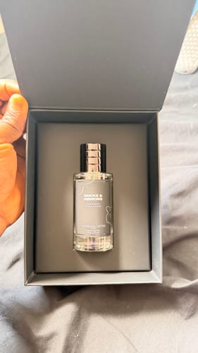 A customer holding the open, dark grey box showing the Smoke & Mirrors Eau de Parfum bottle inside.