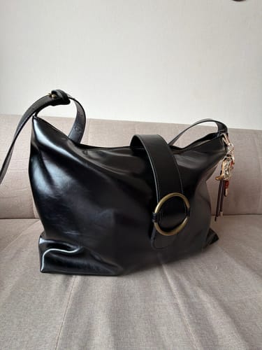 Reviewer's black Cartera De Hombro Argolla Boheme featuring a golden ring detail, resting on a sofa.