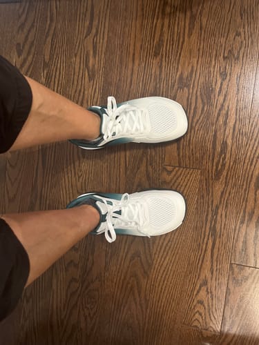 Customer wearing white and teal CUURT Muuv shoes, seen from a top-down view on a dark wood floor.