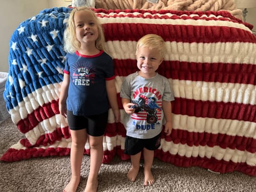 Customer's children posing in front of a soft Hugs Flag Blanket draped over a bed.