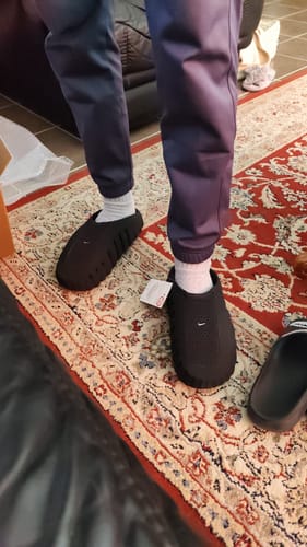 Customer's feet wearing black Nike Mind 001 sneakers with white socks, shown standing on an patterned rug.