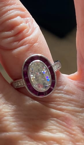Customer's oval moissanite engagement ring with a red ruby halo and milgrain setting on their finger.