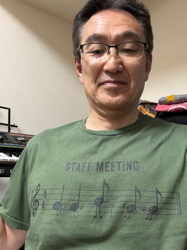 Reviewer wearing a green Staff Meeting 2 T-Shirt with musical note characters, smiling.