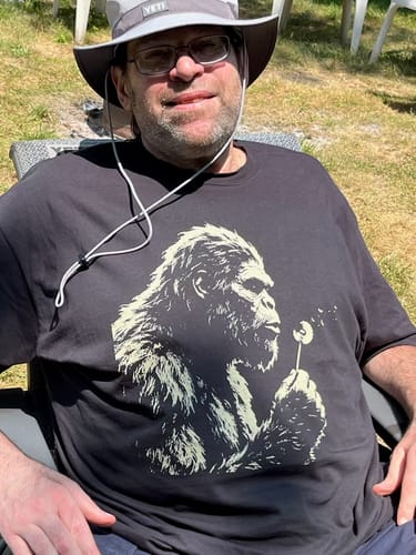 Customer's They Didn't See Us T-Shirt in black with a Sasquatch print, worn outdoors by a person in a sun hat.