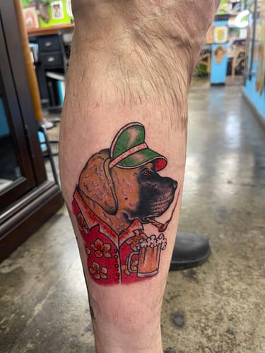 Customer's fresh tattoo of a dog wearing a hat and shirt while holding a beer, done using Signature Tattoo Numbing Cream