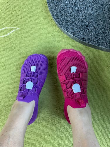 Arvostelijan jalat päällä Vapauta Varpaat™ Paljasjalkakengät, joista toinen on violetti ja toinen kirkkaan vaaleanpunainen.