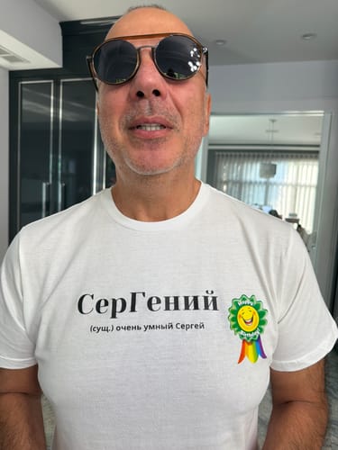 Reviewer wearing a white Sergei Tee featuring a Cyrillic graphic print and a colorful birthday badge.