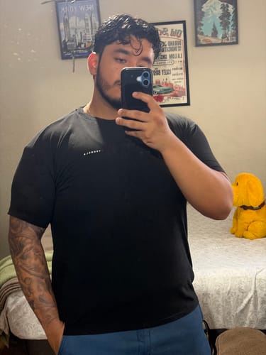 Reviewer taking a mirror selfie to show the snug fit of the black Perfect Tailored Tee on their chest and arms.