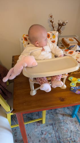 Customer's baby sitting in the Clementine baby chair, which is placed on a wooden table with a toy on the tray.