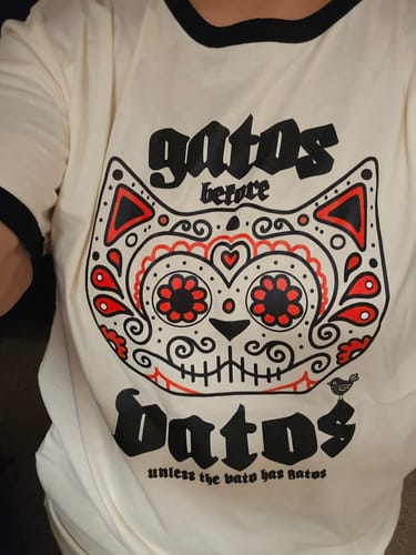 A reviewer's close-up of the Gatos Before Vatos ringer tee, showing the detailed sugar skull cat graphic.