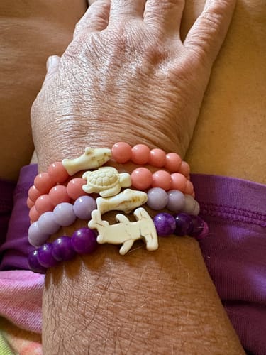 Customer's hand wearing three stacked Shark Week Bracelets with coral, lavender, and purple beads and charms.