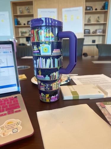 Customer's bookshelf-themed Thicc Buddy mug with purple handle, sitting on a conference table next to a laptop.