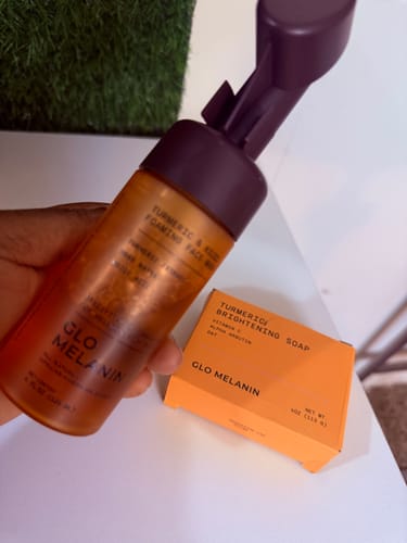 Customer's hand holding a Turmeric Brightening Soap foaming wash bottle next to its bright orange box.