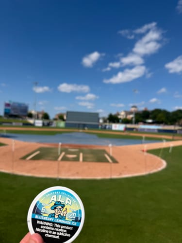 Customer's hand holding a round can of Drifters Blueberry Lemonade Ice nicotine pouches at a sunny baseball field.