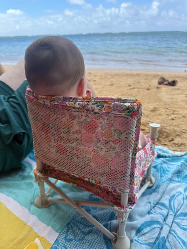 Reviewer's floral print Paloma portable booster chair V2 sitting on a beach towel with a baby facing the ocean.