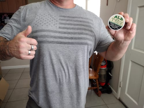 Customer's hand holding a Spearmint Nicotine Pouches container while wearing a grey American flag t-shirt.