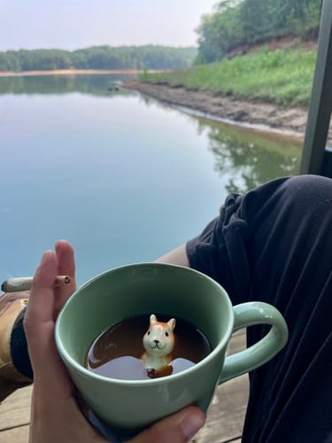 Customer's hand holding a joint of Orange Cookies THCA Flower, sitting by a serene lake with a squirrel mug.