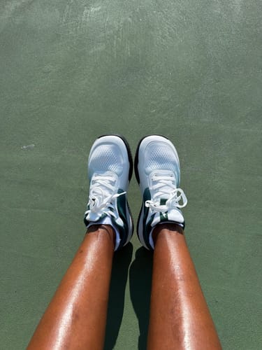 Reviewer's feet wearing white CUURT Muuv court shoes while standing on a green tennis court.