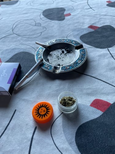 Customer's California Love THC-A flower buds in a small open jar next to rolled cigarettes and a decorative ashtray.