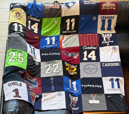 Reviewer's colorful Project Repat T-Shirt Quilt featuring various sports jerseys, laid out on a dark leather couch.