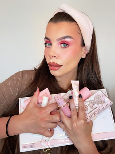 Reviewer holding The Cheek Of It Liquid Blush and beauty products, wearing vibrant pink eye and lip makeup.