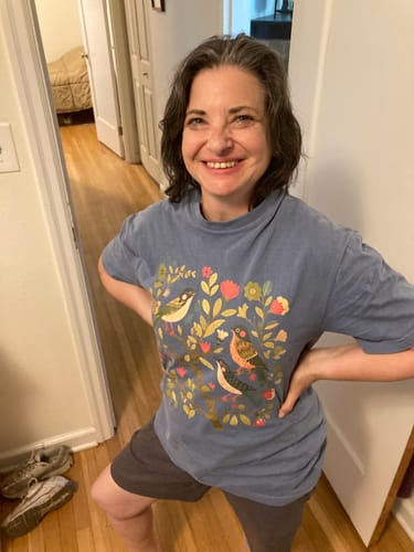 Customer's Cottagecore Birds Folk Art Print T-shirt in blue, worn by a smiling person indoors.