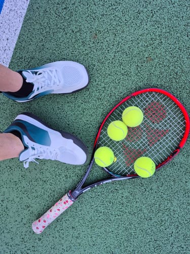 Customer's feet in white CUURT Muuv court shoes on a tennis court next to a racket and balls.