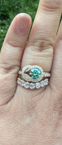 Reviewer's hand showing a custom ring with a teal center stone set in a gold, diamond-paved knot design.